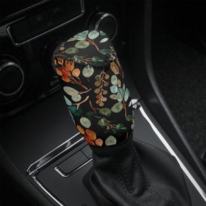 May include: A black gear shift knob with a floral pattern featuring green, orange, and brown leaves. The pattern is on a black background.