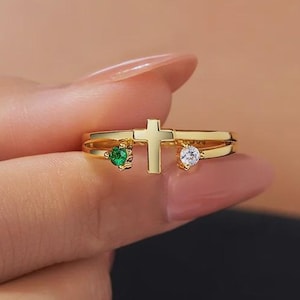Peut inclure: Une bague dorée avec un motif de croix. La bague comporte deux anneaux et est ornée d'une pierre verte et d'une pierre transparente. La bague est tenue par les doigts d'une personne.