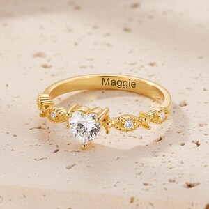 May include: A gold-colored ring with a heart-shaped, clear gemstone at the center. The ring has a delicate design with small, clear stones and the name "Maggie" engraved on the band. The ring is set against a neutral background.