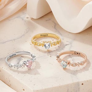 May include: Three personalised rings with heart-shaped gemstones. One is gold with "Maggie" engraved, another is silver with "Soohla", and the third is rose gold with "D K". The rings are displayed on a light-coloured surface.