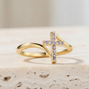 May include: A gold ring with a cross design. The cross is embellished with small, light purple gemstones. The ring has a delicate, curved band. This elegant ring is a spiritual piece of jewellery.