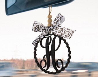 May include: A black wooden car charm with a decorative scalloped circle and the letter "H" in a script font. The charm is adorned with a white and brown leopard print ribbon tied in a bow and a string of wooden beads.