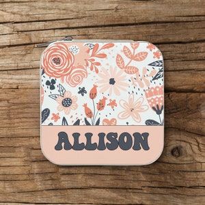 May include: A square, zipped case featuring a floral design in peach and white. The lower section is a solid peach colour with the name "ALLISON" in dark grey, retro lettering. The case is placed on a wooden surface.