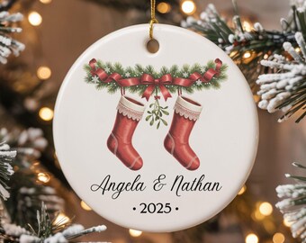 Puede incluir: Adorno navideño redondo y blanco con dos calcetines rojos colgando de una guirnalda de vegetación y cinta roja. Los nombres "Angela & Nathan" y el año "2025" están impresos debajo.