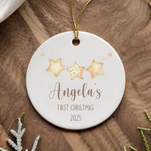 May include: White ceramic Christmas ornament with gold string. The ornament features three watercolor stars and the text "Angela's FIRST CHRISTMAS 2025" in a script font. A festive holiday decoration.