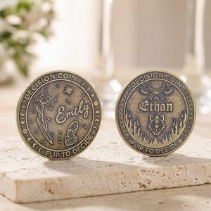 May include: Two circular brass-toned decision coins featuring engraved designs. The left coin displays the name Emily, an iris flower, and an Aquarius symbol. The right coin shows the name Ethan, crossed axes, and a wolf head above flames.