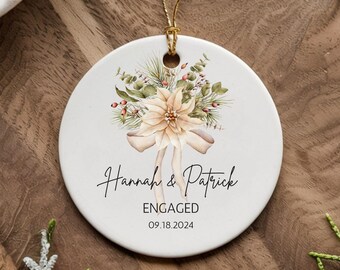 Puede incluir: Un adorno de cerámica blanca con una ilustración de acuarela de una flor de pascua y vegetación. El adorno dice "Hannah & Patrick ENGAGED 09.18.2024".