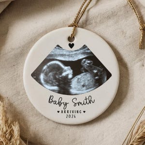 Könnte beinhalten: Runder, cremefarbener Keramikschmuck mit einem Ultraschallbild eines Babys. Der Schmuck hat ein schwarzes Herz und den Text "Baby Smith ♥ ARRIVING ♥ 2026" in schwarzer Schrift. Ein goldener Faden ist zum Aufhängen befestigt.