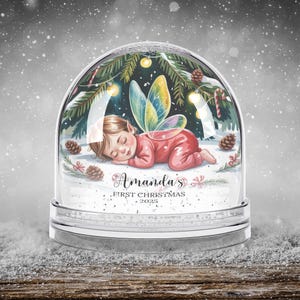 Może przedstawiać: Kula śnieżna z śpiącą wr&oacute;żką w czerwonym stroju i kolorowymi skrzydłami. Kula ma motyw świąteczny z igłami sosnowymi, światłami i napisem "Amanda's First Christmas 2025".