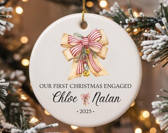 Puede incluir: Adorno navideño redondo de cerámica blanca con un diseño de lazo en acuarela en rojo, dorado y verde. El texto dice "OUR FIRST CHRISTMAS ENGAGED" con los nombres "Chloe" y "Natan" y el año "2025". Cuelga de una cuerda dorada.