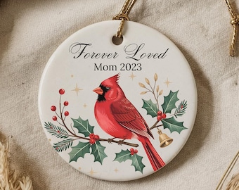 May include: A white ceramic Christmas ornament with a red cardinal bird perched on a holly branch. The ornament has the text "Forever Loved Mom 2023" printed on it.