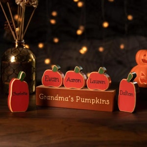 Puede incluir: Cinco pequeñas calabazas de madera con los nombres "Charlotte", "Evan", "Aaron", "Lauren" y "Dalton" pintados en ellas. Las calabazas están sentadas sobre un bloque de madera con el texto "Grandma's Pumpkins" pintado en él.