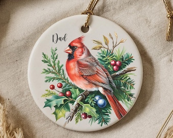 May include: A white ceramic Christmas ornament featuring a red cardinal perched on a branch with green holly and berries. The ornament is round and has a gold string for hanging. The word "Dad" is written on the ornament.