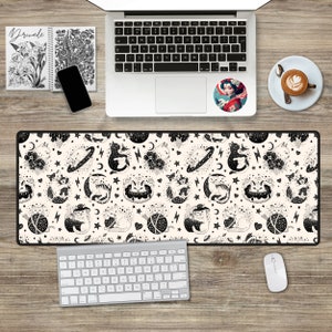 May include: A black and white desk mat featuring a repeating pattern of cats, stars, moons, and other celestial objects. The mat is on a wooden desk with a laptop, keyboard, mouse, and a cup of coffee.