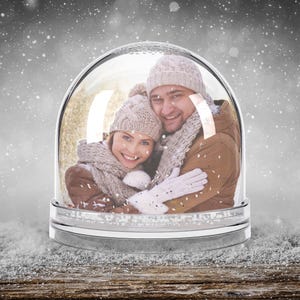 Könnte beinhalten: Eine Schneekugel mit einem Paar darin, die Wintermützen und Schals tragen. Die Kugel steht auf einer Holzoberfläche mit einem verschneiten Hintergrund.