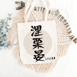 May include: A white canvas tote bag with black Chinese characters and a geometric pattern in a circle. The text "LILIAN" is printed below the circle.