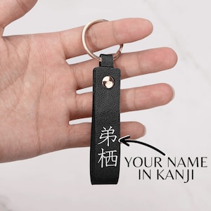 May include: Black leather keychain with a gold metal ring. The keychain has a white inscription in Japanese characters, "久保".