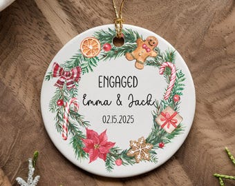 Puede incluir: Adorno navideño de cerámica blanca con un diseño de corona verde. La corona está decorada con bastones de caramelo rojo y blanco, un hombre de jengibre, una flor de pascua roja y un lazo rojo. El adorno dice "ENGAGED Emma & Jack 02.15.2025".