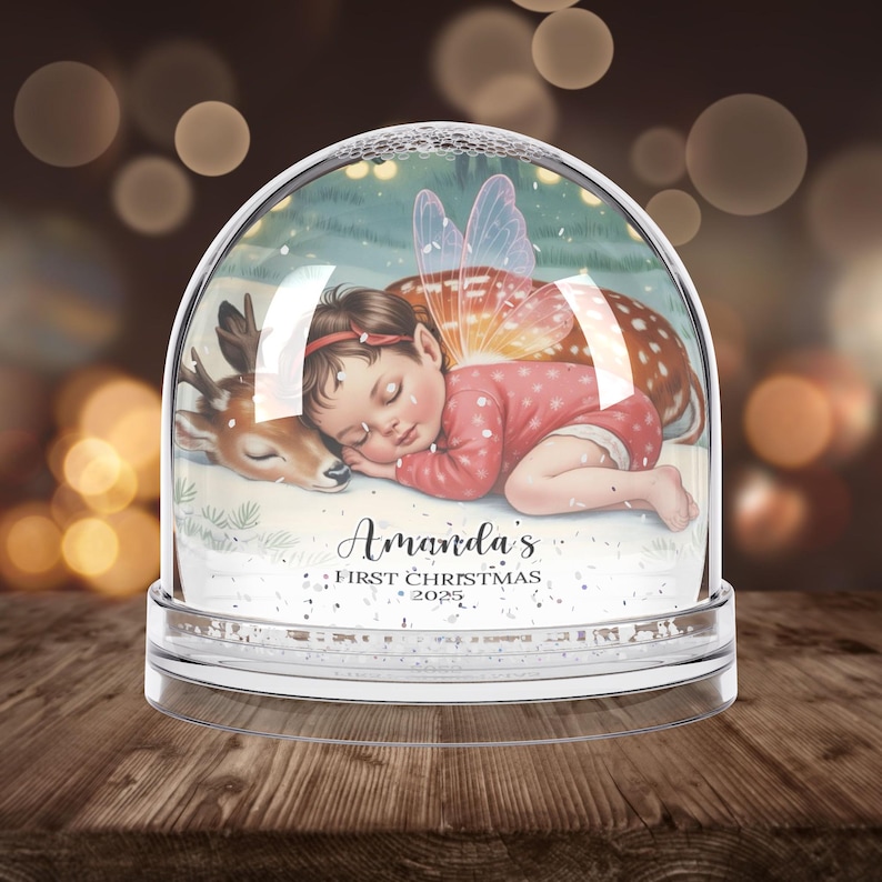 K&ouml;nnte beinhalten: Eine klare Schneekugel mit einem schlafenden Baby-Fee und einem Hirsch darin. Das Baby tr&auml;gt einen rosa Anzug und hat Feenfl&uuml;gel. Auf der Basis steht der Text "Amanda's First Christmas 2025".