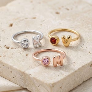 May include: Three open-style rings featuring a single letter and a round gemstone. One silver-tone ring has a clear stone and the letter K, one gold-tone ring has a red stone and the letter V, and one rose gold-tone ring has a pink stone and the letter M.