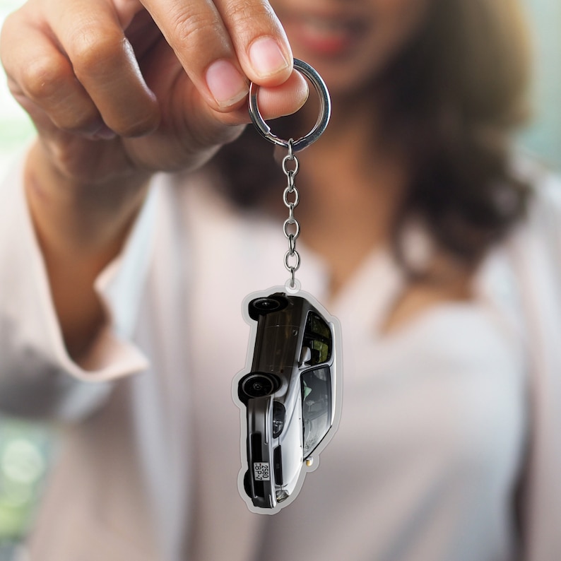 May include: A silver keychain with a clear plastic charm featuring a gray car. The car is upside down with the license plate visible.