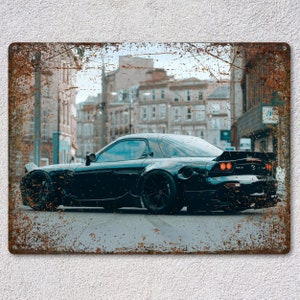 May include: A black sports car with a rear spoiler is parked on a city street. The image is printed on a metal sign with a distressed, rusty finish. The background features buildings and a cloudy sky.