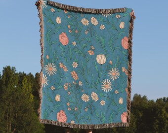 Puede incluir: Una manta turquesa con un estampado floral de margaritas, tulipanes y otras flores en tonos melocotón, crema y verde. La manta tiene un borde con flecos y se sostiene contra un cielo azul.