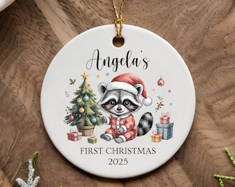Può includere: Ornamento natalizio in ceramica bianca con un procione con cappello da Babbo Natale e abito rosso, un albero di Natale e regali. Il testo "Angela's First Christmas 2025" è stampato sull'ornamento.