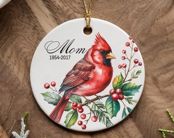 May include: White ceramic ornament with a red cardinal perched on a branch with red berries and green leaves. The ornament has the text "Mom 1954-2017" printed on it.