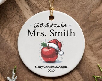 May include: A white ceramic Christmas ornament with a gold string. The ornament features the text "To the best teacher Mrs. Smith" and a red apple wearing a Santa hat. The text "Merry Christmas, Angela 2025" is also included.