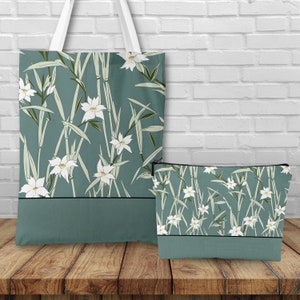 May include: A teal tote bag and matching cosmetic bag with a white floral pattern on a white brick background. The floral pattern features white flowers and green leaves.