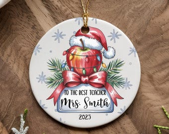 May include: A round, white Christmas ornament with a festive design. It features a red apple wearing a Santa hat and adorned with lights, a red bow, and the text "TO THE BEST TEACHER Mrs. Smith 2023". The ornament has a gold string for hanging.