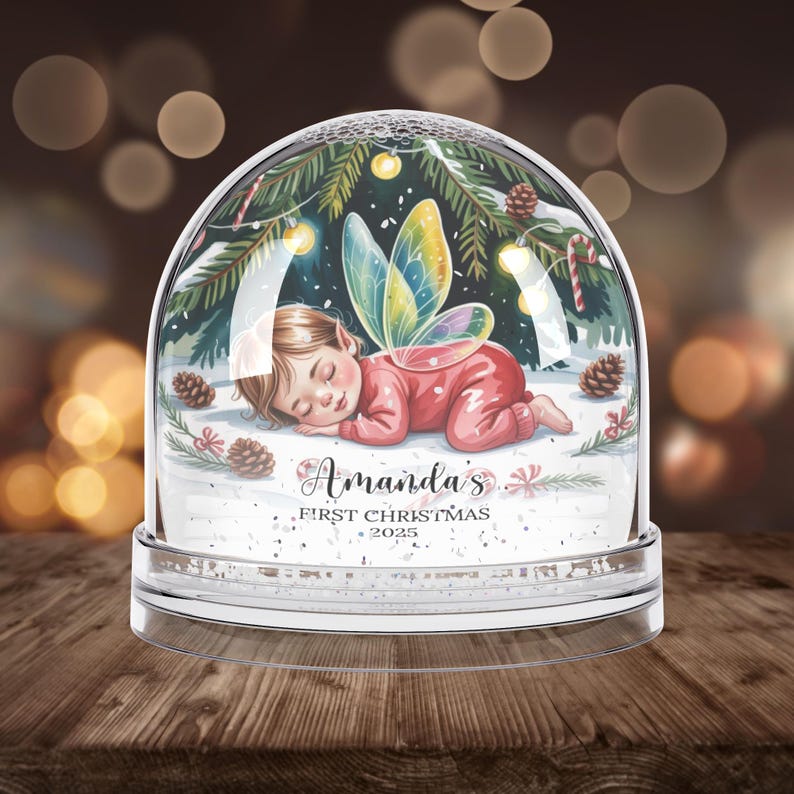 Może przedstawiać: Przezroczysta szklana kula śnieżna ze śpiącą wr&oacute;żką w środku. Dziecko ma na sobie czerwony str&oacute;j i kolorowe skrzydła. Kula jest ozdobiona igłami sosnowymi, światełkami i napisem "Amanda's First Christmas 2025."