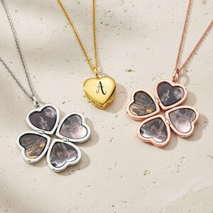 Peut inclure: Trois colliers avec des pendentifs en forme de cœur. L'un est argenté avec un design de trèfle à quatre feuilles, chaque feuille contenant une photo. Un autre est doré avec la lettre "A". Le troisième est en or rose, également avec un design de trèfle à quatre feuilles.