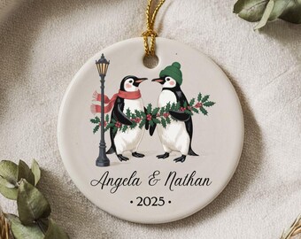May include: A round, cream-colored Christmas ornament featuring two penguins. One penguin wears a red scarf, the other a green hat. They hold a holly branch. A black lamppost is on the left. The names "Angela & Nathan" and the year "2025" are printed below.