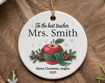 May include: A white ceramic Christmas ornament with a gold string. The ornament features an illustration of a red apple wrapped in a green scarf, with text that reads "To the best teacher Mrs. Smith" and "Merry Christmas, Angela 2025".