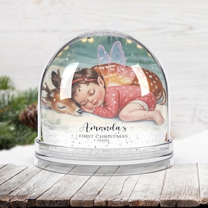 K&ouml;nnte beinhalten: Eine klare Schneekugel mit einem schlafenden Baby-Fee und einem Rehkitz darin. Das Baby tr&auml;gt einen rosa Anzug und hat Feenfl&uuml;gel. Der Text "Amanda's First Christmas 2025" ist unten geschrieben.