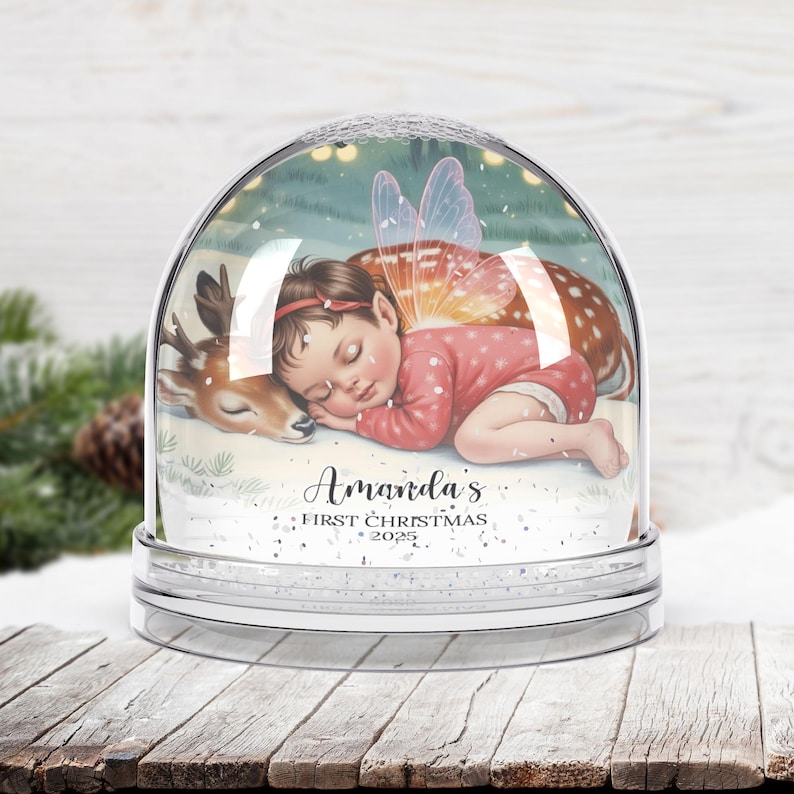 K&ouml;nnte beinhalten: Eine klare Schneekugel mit einem schlafenden Baby-Fee und einem Rehkitz darin. Das Baby tr&auml;gt einen rosa Anzug und hat Feenfl&uuml;gel. Der Text "Amanda's First Christmas 2025" ist unten geschrieben.