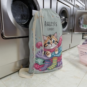 May include: A light blue laundry bag with a drawstring closure. The bag features a cartoon kitten design with the text "Ashley's Laundry". The kitten is surrounded by colorful socks and clothing.