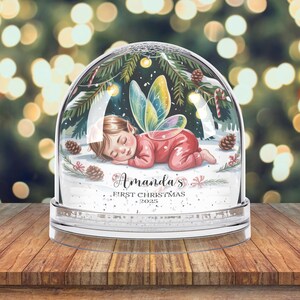 Może przedstawiać: Przezroczysta szklana kula śnieżna ze śpiącą wr&oacute;żką w czerwonym stroju i kolorowymi skrzydłami. Na podstawie znajduje się napis "Amanda's First Christmas 2025". Tło przedstawia rozmyte światła świąteczne, tworząc świąteczną atmosferę.