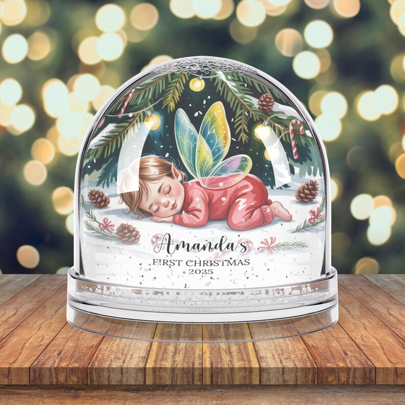 Może przedstawiać: Przezroczysta szklana kula śnieżna ze śpiącą wr&oacute;żką w czerwonym stroju i kolorowymi skrzydłami. Na podstawie znajduje się napis "Amanda's First Christmas 2025". Tło przedstawia rozmyte światła świąteczne, tworząc świąteczną atmosferę.