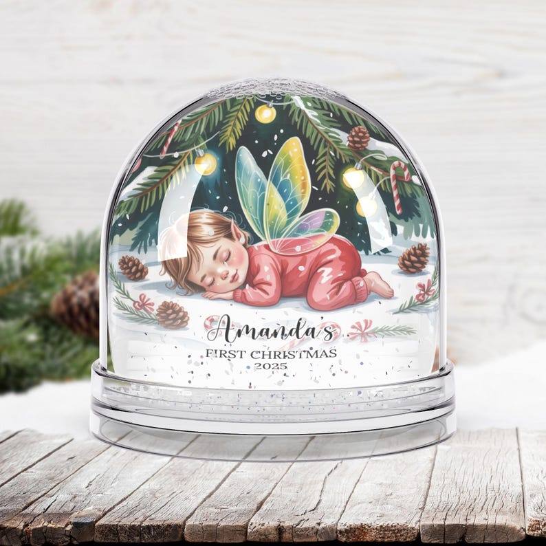 Może przedstawiać: Przezroczysta kula śnieżna z śpiącą wr&oacute;żką w czerwonym stroju i kolorowymi skrzydłami. Kula jest ozdobiona igłami sosnowymi, cukrowymi laskami i napisem "Amanda's First Christmas 2025". Podstawa jest przezroczysta.