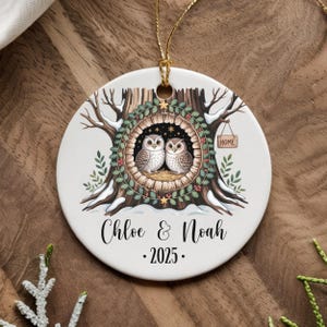 May include: A round ceramic Christmas ornament featuring two owls nestled in a tree hollow. The ornament has a gold hanging cord and includes the names "Chloe & Noah" and the year "2025". The design incorporates winter elements.