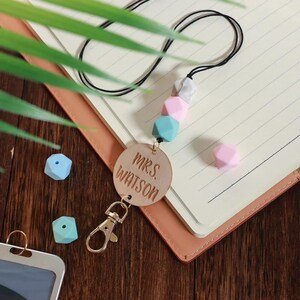 以下が含まれることがあります： 「MRS. WATSON」と刻まれた茶色の革製のキーホルダー。キーホルダーには金色の金属製のクリップと、カラフルなシリコンビーズが付いた黒いコードが付いています。