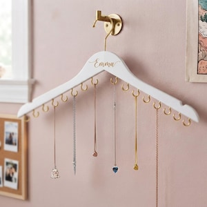 May include: A white wooden clothes hanger mounted on a gold wall hook, featuring the name Emma in gold script. The hanger includes twelve gold hooks along the bottom edge, with several delicate metal necklaces hanging from the hooks.