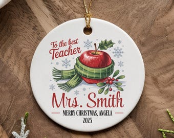 May include: A white ceramic Christmas ornament with a gold string. The ornament features a red apple wearing a green scarf, snowflakes, and the text "To the best Teacher Mrs. Smith Merry Christmas, Angela 2025."