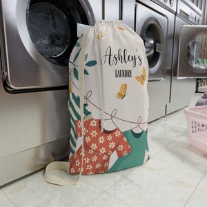 May include: A white laundry bag with a drawstring closure, featuring the text "Ashley's LAUNDRY" in black script. The bag is decorated with butterflies, floral and clothing illustrations. A beige strap is attached to the bag.