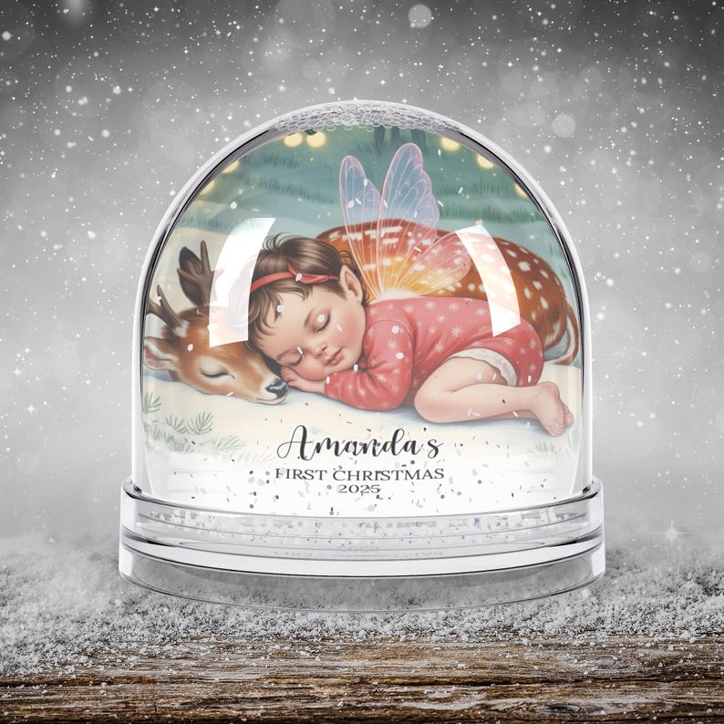 K&ouml;nnte beinhalten: Eine Schneekugel mit einem schlafenden Baby-Fee und einem Reh. Das Baby tr&auml;gt ein rosa Outfit und hat Feenfl&uuml;gel. Der Text "Amanda's First Christmas 2025" steht auf der Basis der Schneekugel.