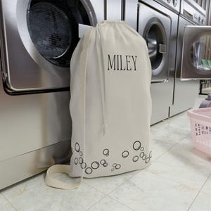 May include: A white canvas laundry bag with black drawstring and black bubbles around the bottom. The bag has the name "MILEY" printed in black on the front.