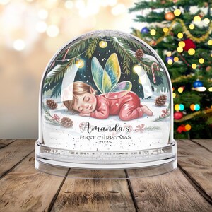 Może przedstawiać: Przezroczysta kula śnieżna ze śpiącą wr&oacute;żką w środku. Dziecko ma na sobie czerwony str&oacute;j i kolorowe skrzydła. Kula jest ozdobiona igłami sosnowymi, śniegiem i napisem "Amanda's First Christmas 2025".